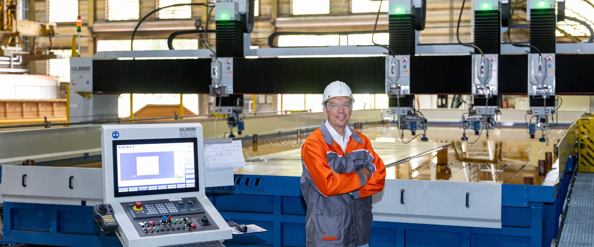 Portrait Robert Kühn vor Wasserstrahlschneidanlage Robert Kühn steht mit verschränkten Armen in einer Werkhalle vor einer großformatigen Wasserstrahlschneidanlage. Im Vordergrund sind das Bedienpult und der Bildschirm zu sehen; im Hintergrund befinden sich mehrere Schneidköpfe der Anlage.