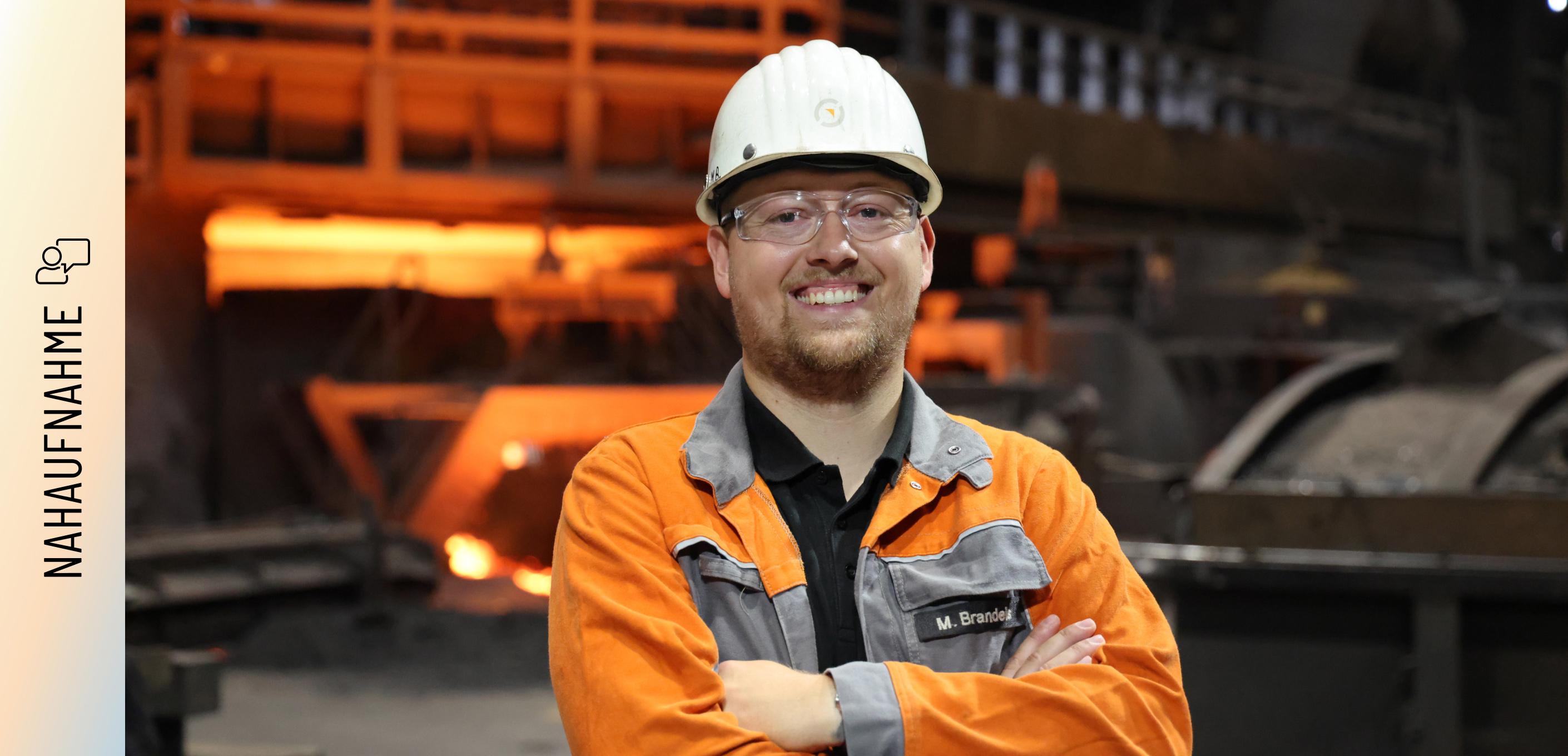 Porträt von Marvin Brandeis Marvin Brandeis steht mit verschränkten Armen und einem Lächeln vor einem Hochofen. Er trägt eine orangefarbene Arbeitsschutzjacke mit seinem Namenszug, einen weißen Schutzhelm und eine Schutzbrille. Im Hintergrund glüht das rötliche Licht der Produktionsanlage.